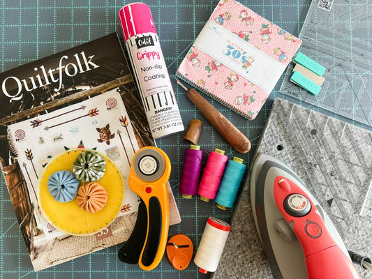 A collection of quilting supplies such as thread, iron, rotary cutter, seam ripper, and a copy of Quiltfolk magazine