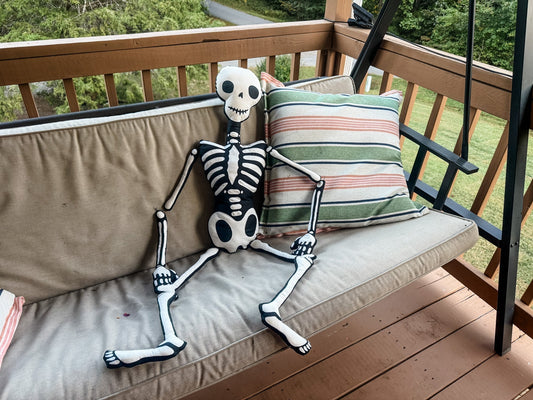 A stuffed skeleton toy sitting on a porch swing next to a striped pillow